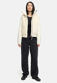 Crème-kleurige pufferjack met een zachte fleece body, hoge kraag, volledige rits en geribde manchetten, gecombineerd met zwarte baggy jeans en zwarte sneakers.