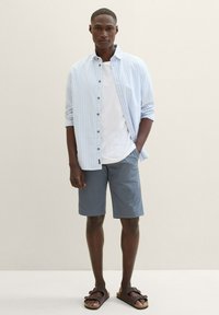 Hellblau-weiß gestreiftes Hemd über einem weißen T-Shirt, kombiniert mit grauen Shorts und braunen Sandalen.