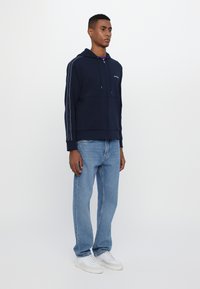 Felpa con zip navy, dotata di cordoncino, strisce laterali e tasche frontali, abbinata a jeans azzurri e sneakers bianche a bassa altezza.