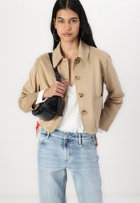 Veste beige courte avec une texture lisse, ornée de boutons ronds et d'une poche poitrine. Associée à un jean clair et un petit sac noir.