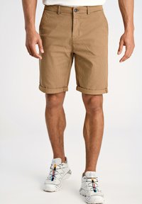 Shorts en velours côtelé marron avec ourlets roulés, dotés d'une fermeture à bouton et de poches avant, portés avec des chaussures de sport grises.