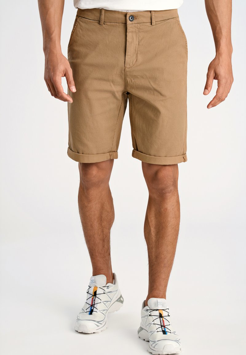Shorts en velours côtelé marron avec ourlets roulés, dotés d'une fermeture à bouton et de poches avant, portés avec des chaussures de sport grises.