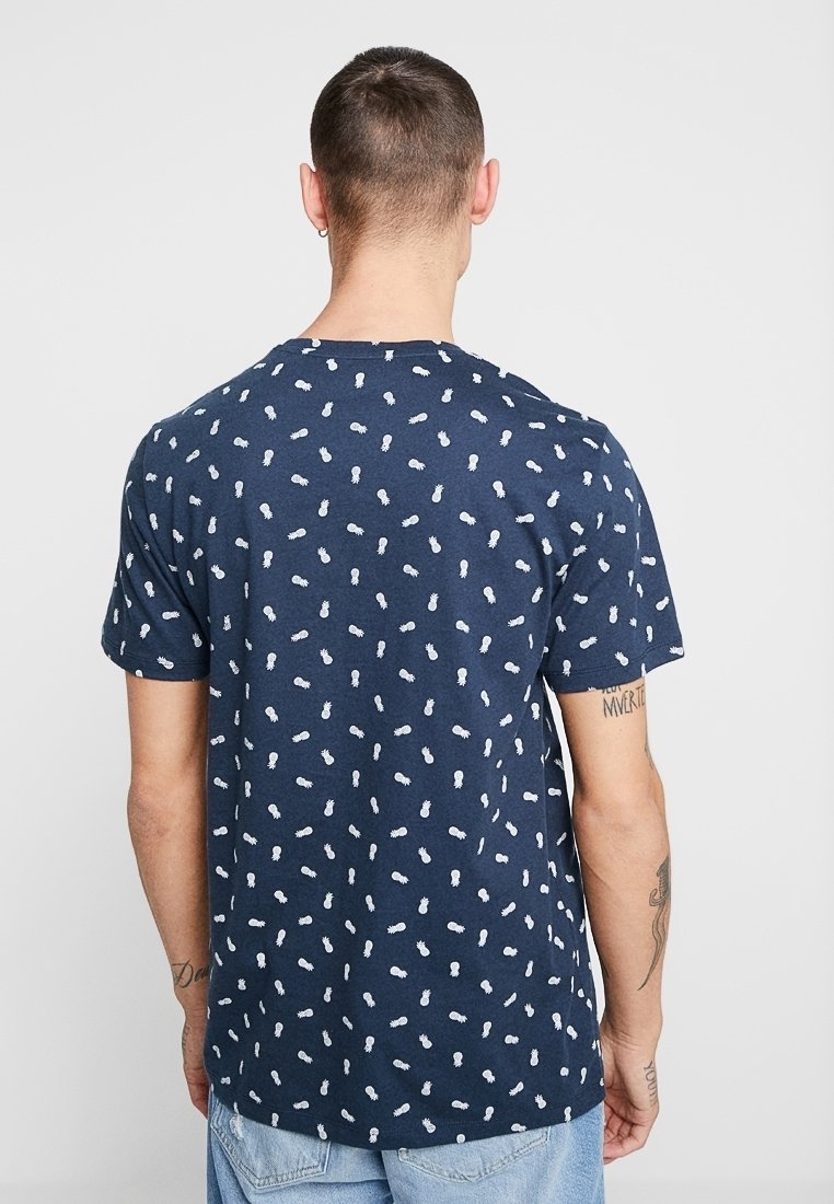 Camiseta de algodón azul marino con mangas cortas, con un patrón disperso de piñas blancas por todo el tejido. Cuello plano, corte casual.