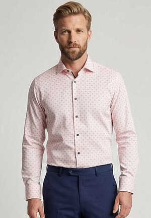 Homme aux cheveux blonds et à la barbe portant une chemise rose clair à manches longues avec de petits pois et un pantalon bleu foncé sur un fond uni.