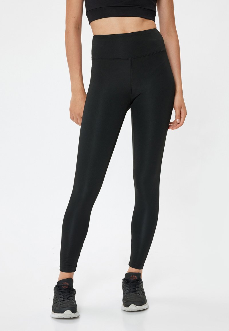 Leggings noirs taille haute en tissu extensible, avec une texture lisse et un design ajusté, associés à des chaussures de sport sombres.