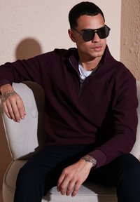 Suéter burdeos con cremallera, textura acanalada, cuello con solapa y mangas largas, combinado con pantalones oscuros. Modelo con gafas de sol negras.