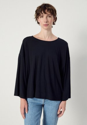 Persona con capelli corti e mossi che indossa una maglia nera larga a maniche lunghe e jeans azzurri chiari, in piedi davanti a uno sfondo chiaro uniforme.