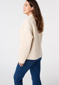 Beige gewatteerde jas met een textuurpatroon, ronde kraag, lange mouwen en een losse pasvorm, gedragen met blauwe spijkerbroeken.