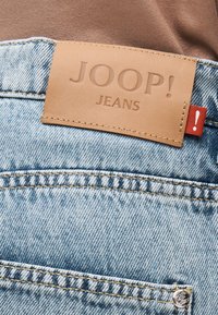 Ανοιχτό μπλε τζιν παντελόνι με ταμπόν από δέρμα σε αποχρώσεις καφέ με την επιγραφή «JOOP! JEANS» και μια μικρή κόκκινη υφασμάτινη ετικέτα με θαυμαστικό.