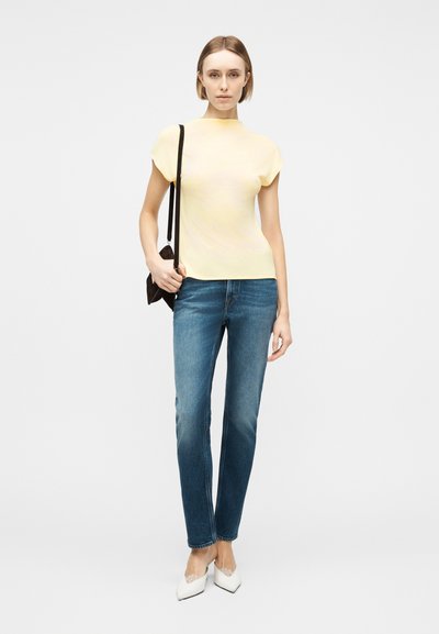 Γυναίκα που φοράει κοντομάνικη μπλούζα ανοιχτού κίτρινου χρώματος, μπλε τζιν, λευκά μυτερά παπούτσια και κρατάει μαύρη τσάντα ώμου, στέκεται μπροστά σε λευκό φόντο.