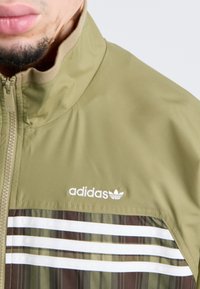 Maslena olivna Adidas jakna s visokim ovratnikom, zipperskim zatvaranjem i horizontalnimi belimi črtami na vzorčnem rjavem delu.