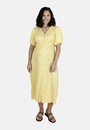 Vrouw staat, glimlacht, draagt een gele midijurk met witte bloemenprint en bruine sandaaltjes met bandjes, tegen een witte achtergrond.