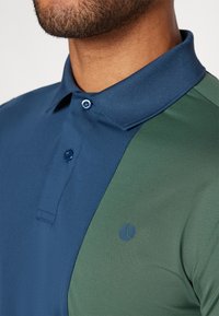 Polo särk, millel on sinine vasak pool ja tumeroheline parem pool. Tekstuuriga kangas, krae ja väike logo rinnal.