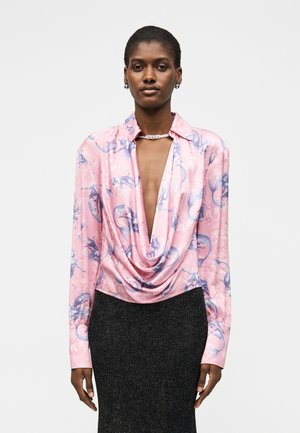 Vrouw draagt een roze satijnen blouse met blauwe draakprints, diepe gedrapeerde halslijn, gouden kettingketting en een donkere gestructureerde rok, staand in een neutrale houding.