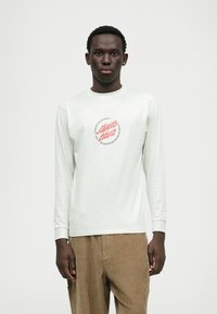 Long-sleeve light gray cotton shirt with a circular red graphic design on the chest. Corduroy beige pants with a textured surface.