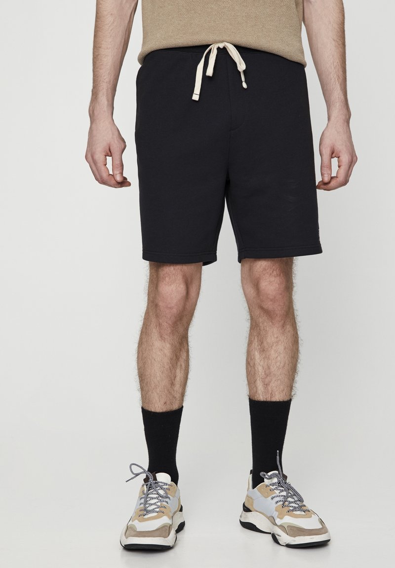 Shorts noirs en coton, avec une taille élastique et un cordon de serrage beige. Portés avec un haut beige et des baskets.