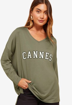 Olijfgroene langemouwen V-hals top met "CANNES" in vette witte letters. Zachte stof en een ontspannen pasvorm met zijsplitten voor extra comfort.
