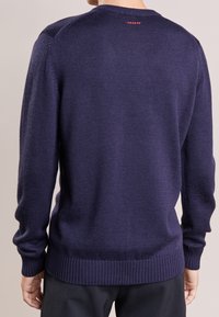 Marineblauwe gebreide trui met een geribde rand en manchetten, met een klein rood logo detail op de achterkant van de halslijn. Glad textuur en standaard pasvorm.