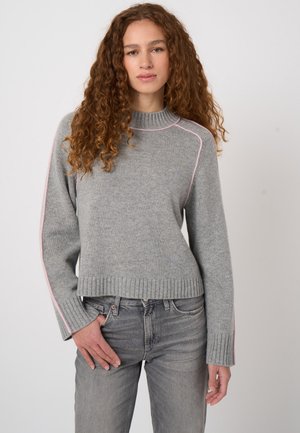 Junge Frau mit langen lockigen Haaren, die einen grauen Pullover mit pinken Nähten und graue Jeans trägt, steht vor einem schlichten Hintergrund.