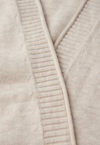 Beige kofta med en mjuk textur, med V-ringning och ribbade detaljer längs kanterna. Tyget har ett slätt och mysigt utseende.