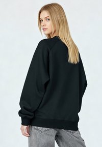 Sweat-shirt noir à coupe décontractée, avec manches raglan et poignets côtelés, associé à un jean gris clair. Le mannequin a les cheveux blonds longs.