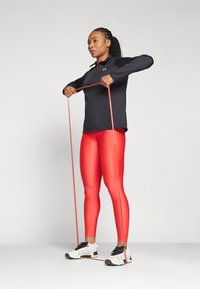 Haut de sport noir à manches longues, leggings rouges et baskets blanches. Individu étirant une bande de résistance rouge, posture concentrée, arrière-plan neutre.