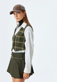 Grønternet vest over en hvid skjorte, parret med en mørkegrøn stribet nederdel og en brun læderkasket. Outfitet inkluderer en bælte detalje.