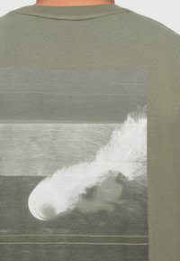 Oliwkowa zielona koszulka z szarym abstrakcyjnym wzorem fali na plecach, z teksturowanymi wzorami i gładkim materiałem.