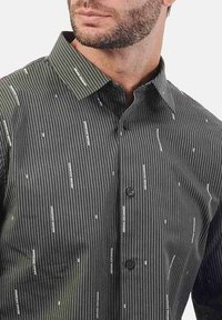 Camicia da uomo con bottoni di colore verde scuro, con righe verticali bianche e motivo texture. Presenta un colletto a contrasto e due bottoni frontali.