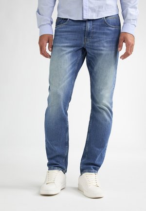 Hombre con vaqueros azules desgastados, zapatillas blancas y una camisa de manga larga azul claro, de pie contra un fondo liso.