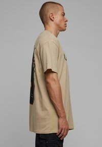Beige cotton t-shirt with a loose fit, crew neckline, short sleeves, and a graphic design printed on the back. Side view shows a straight silhouette.