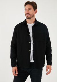 Camisa negra de botones hecha de tela texturizada, con un bolsillo frontal, mangas largas y un dobladillo curvado, llevada sobre una camiseta blanca.