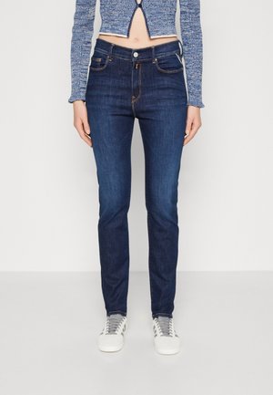 Skinny dark blue jeans made of denim with a faded finish. Features five pockets, orange stitching, and a buttoned closure. Paired with gray sneakers.