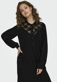 Czarna sukienka z długim rękawem, z koronkową wstawką przy dekolcie, z guzikami z przodu oraz z marszczonymi mankietami, wykonana z lekkiego materiału.