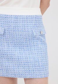 Sininen tweed mini-hame, jossa on teksturoitu ruutukuosi, valkoinen vyötärö ja kaksi etupussia hopeanvärisillä yksityiskohdilla.