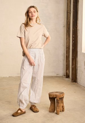 Frau in beigem T-Shirt, weißen gestreiften weiten Hosen und braunen Schuhen steht neben einem kleinen rustikalen Holzschemel in einem hellen, kahlen Raum.