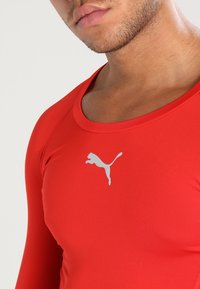 Rød langermet treningstopp laget av glatt, elastisk stoff med rund hals og en grå Puma-logo på brystet.