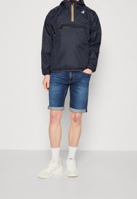 Man wearing navy windbreaker with yellow zipper, rolled-up denim shorts, white socks, and white Diesel sneakers standing on a plain background.