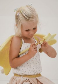 Dieťa s blond vlasmi v bielom kvietkovanom topi a sukni s zlatými akcentmi. Drží žltú paličku s hebkými detailmi hviezdy a brmbolca a má na sebe žlté krídla.