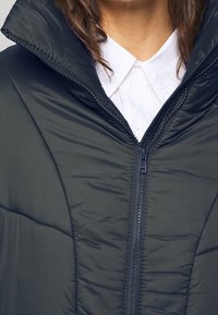 Giacca a vento navy con colletto alto, chiusura zip e texture trapuntata. Indossata sopra una camicia bianca con bottoni, caratterizzata da un design elegante.