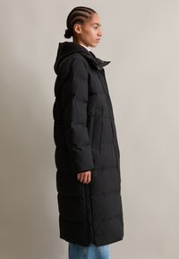 Vrouw in zijaanzicht, gekleed in een lange zwarte gewatteerde jas met capuchon, blauwe jeans en een wit topje voor een effen beige achtergrond.