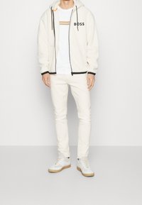 Weißer Fleecepullover mit Reißverschluss und schwarzen Akzenten, weißes T-Shirt mit beigen Streifen, cremefarbene Slim-Fit-Jeans und weiße Turnschuhe mit Gummisohlen.