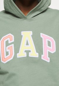 Sudadera con capucha de algodón verde con la palabra "GAP" en letras grandes bordadas: rosa, amarillo y naranja con contorno blanco. Textura suave.