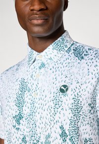 Short-sleeve button-up shirt in white with green floral pattern; features a collar and a round logo patch. Textured fabric.