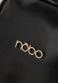 Musta synteettinen laukku, jossa on liukas rakenne ja kultasävyinen "nóbo"-logo. Logo on kohokuvioitu, mikä luo kontrastia pinnan kanssa.