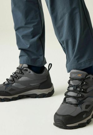 Primer plano de pies con zapatos de senderismo Regatta grises y negros y pantalones azul oscuro sobre un fondo claro.