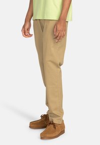 Chino en coton beige avec une coupe droite, texture légère et poches, associés à des chaussures en daim marron avec des lacets.