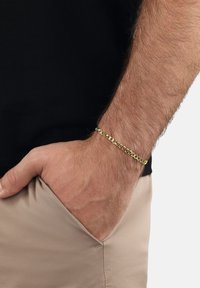 Pulsera de cadena de oro con eslabones entrelazados, llevada en una muñeca, presenta un acabado liso y una superficie pulida. El fondo es neutral.