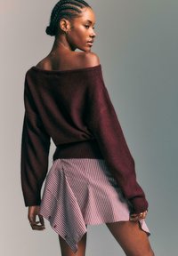 Bordo oversize pulover z teksturirano tkanino in obliko brez ramen, v kombinaciji z rožnato in belo prugasto asimetrično krilo.