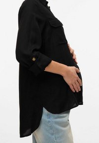 Blusa negra de manga larga con dos bolsillos en el pecho, dobladillo redondeado y detalles de botones dorados en los puños, usada sobre jeans de denim claros.